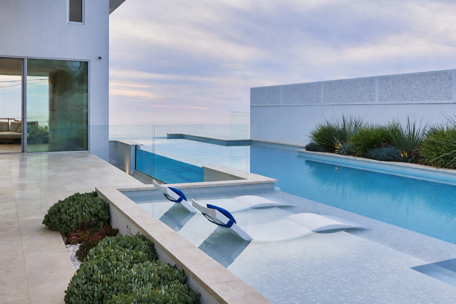 Modern white coastal residence at dusk with multi-level pool featuring turquoise acrylic window panels and ocean backdrop